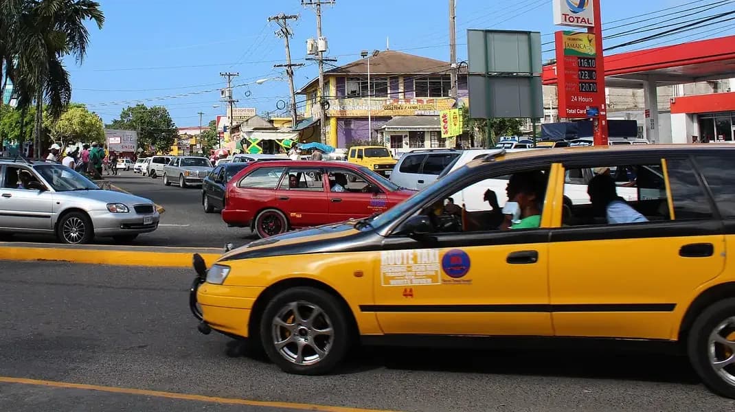 A yellow route taxi with passengers on a Kingston street, shopfronts, a Jamaican flag and palms behind it